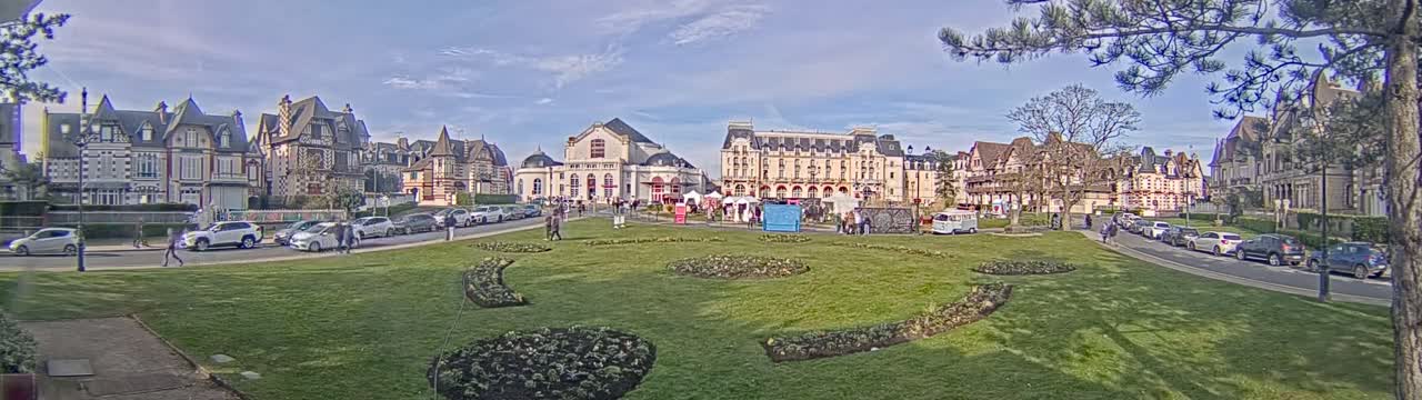 A grassy park with flowerbeds is situated between rows of parked cars and a collection of buildings under a mostly sunny sky.