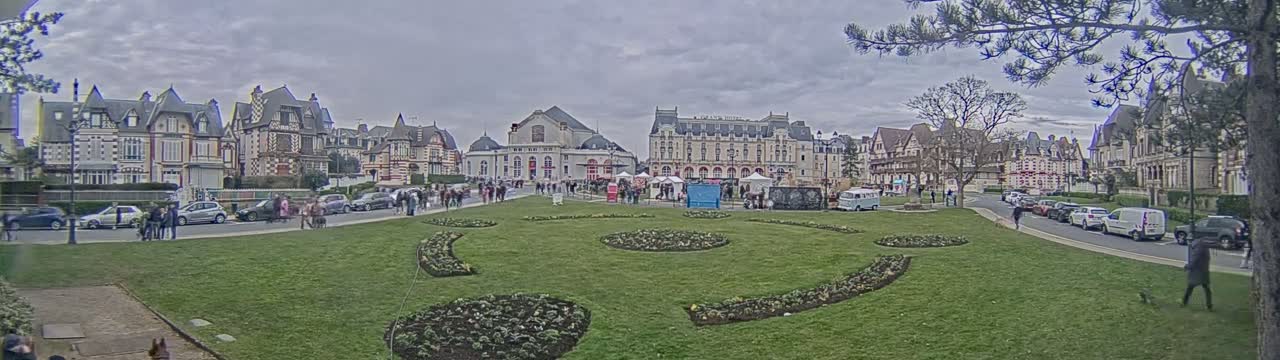 A town square with ornate buildings and a grassy area with flowerbeds, under an overcast sky.