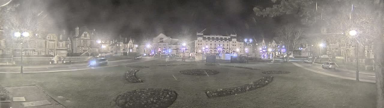 A nighttime scene shows several buildings, cars, and streetlights in a town square with landscaped areas, possibly experiencing light snow or rain.