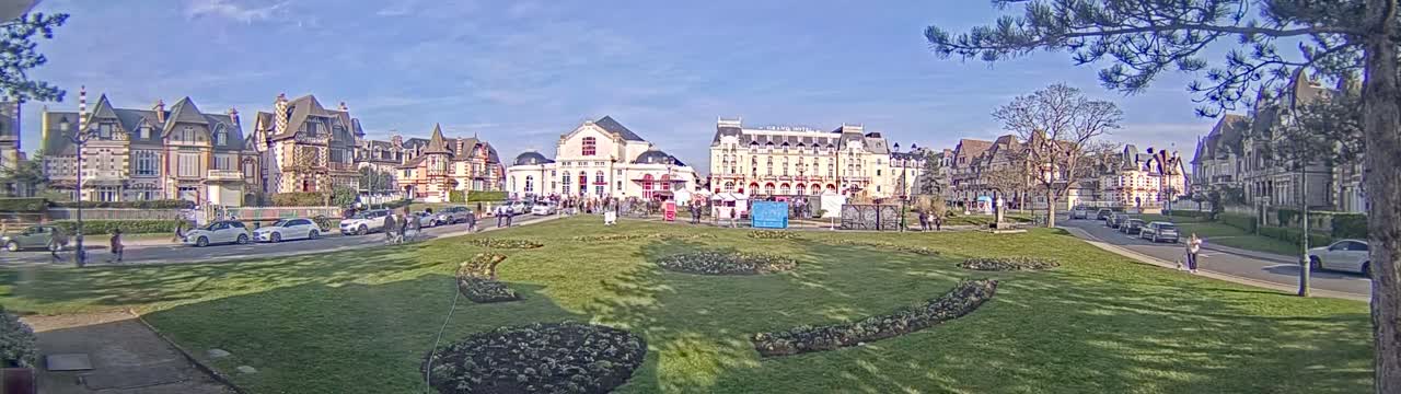 A sunny day reveals a grassy town square with several ornate buildings and parked cars, surrounded by trees and pedestrians.