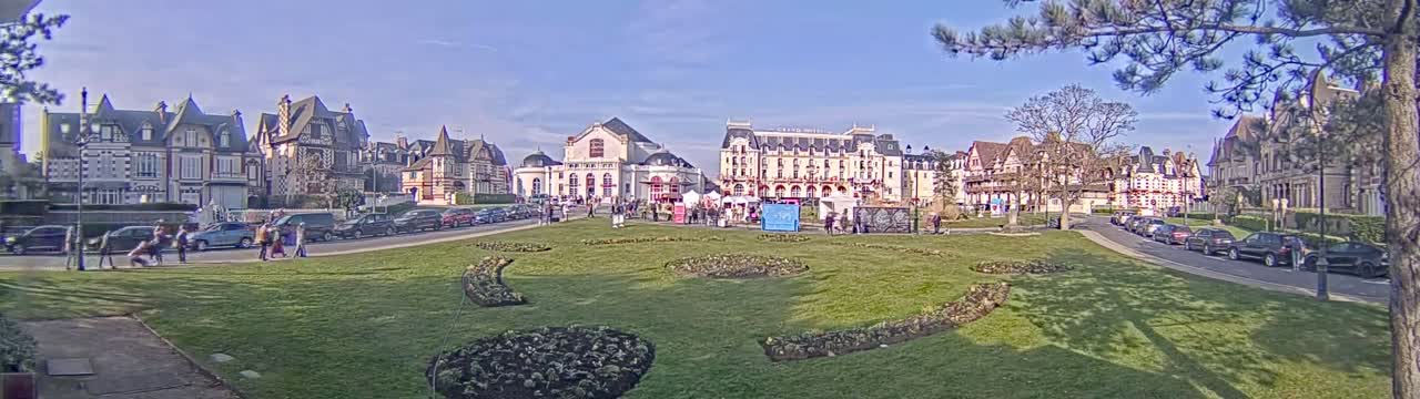 A sunny day reveals a town square with several large buildings, parked cars, and pedestrians near manicured lawns and flowerbeds.