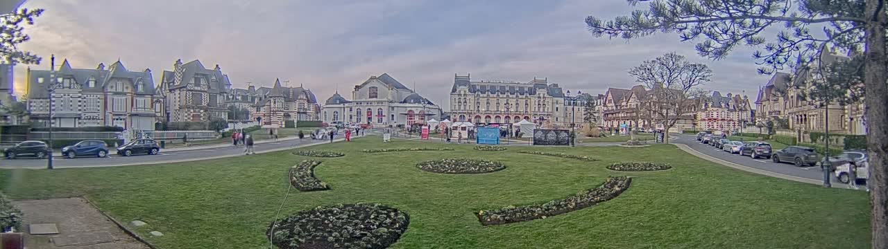 A town square with several large buildings and manicured lawns under an overcast sky, with cars parked along the streets and a few people walking around.