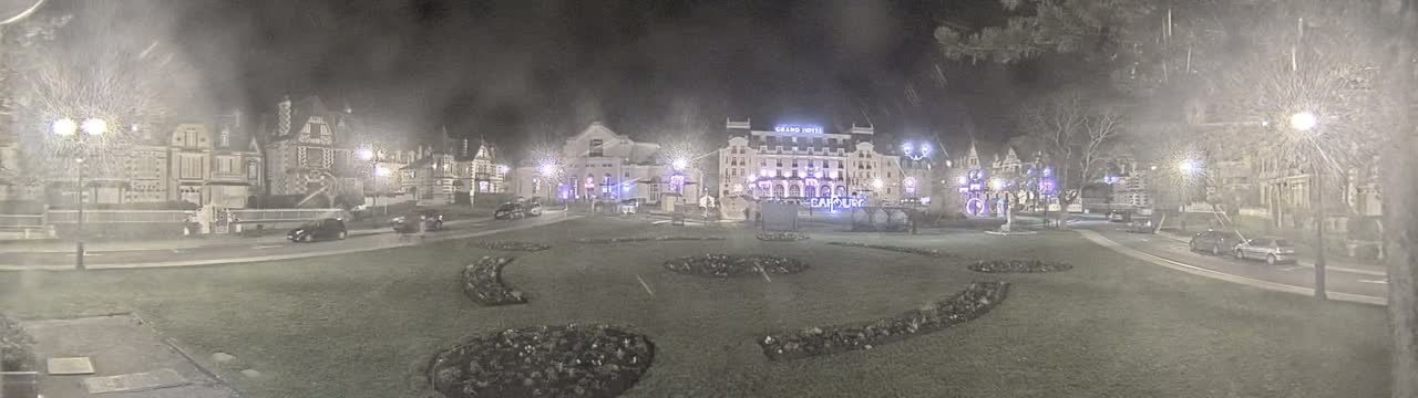 A nighttime view of a town square with several buildings, including a large hotel, with light snow or mist falling.