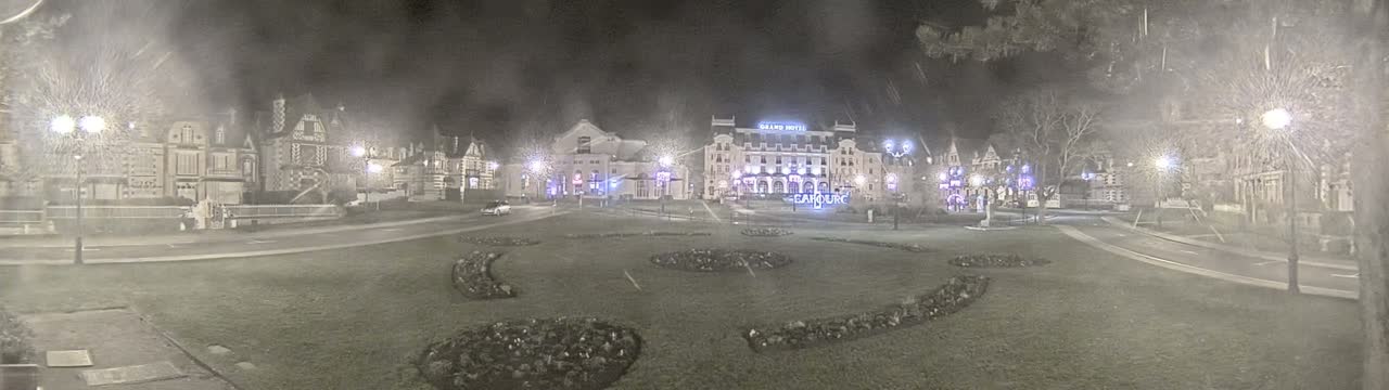 A nighttime view of a town square with several buildings, including a large hotel, with some decorative landscaping and streetlights under hazy or snowy conditions.