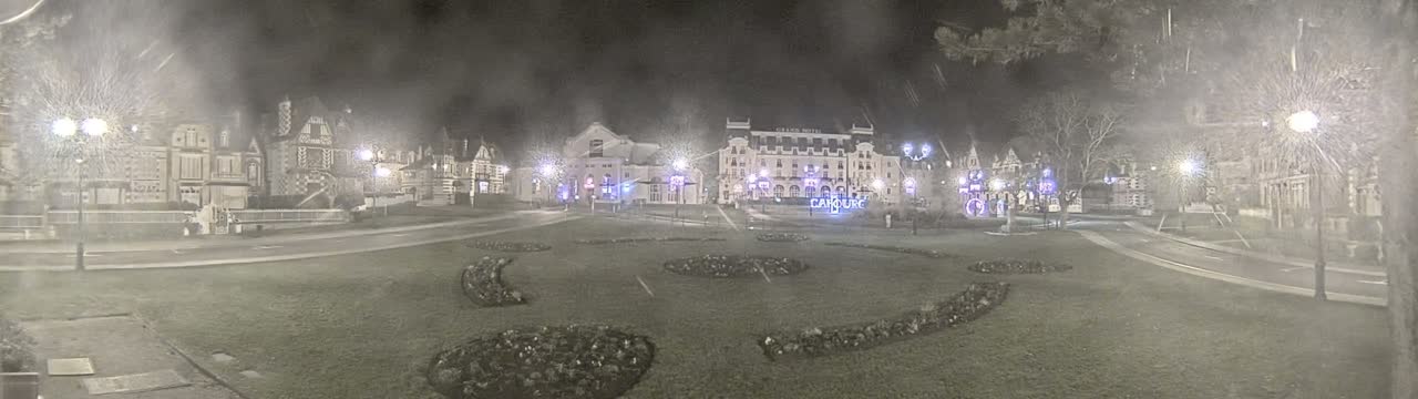 A nighttime view of a town square with several buildings, including a large hotel, surrounded by a grassy area with landscaped flowerbeds under hazy or snowy conditions.