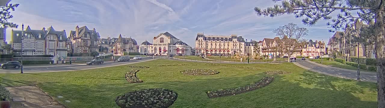 A row of large, ornate buildings sits across from a grassy park with landscaped flowerbeds under a mostly sunny sky.