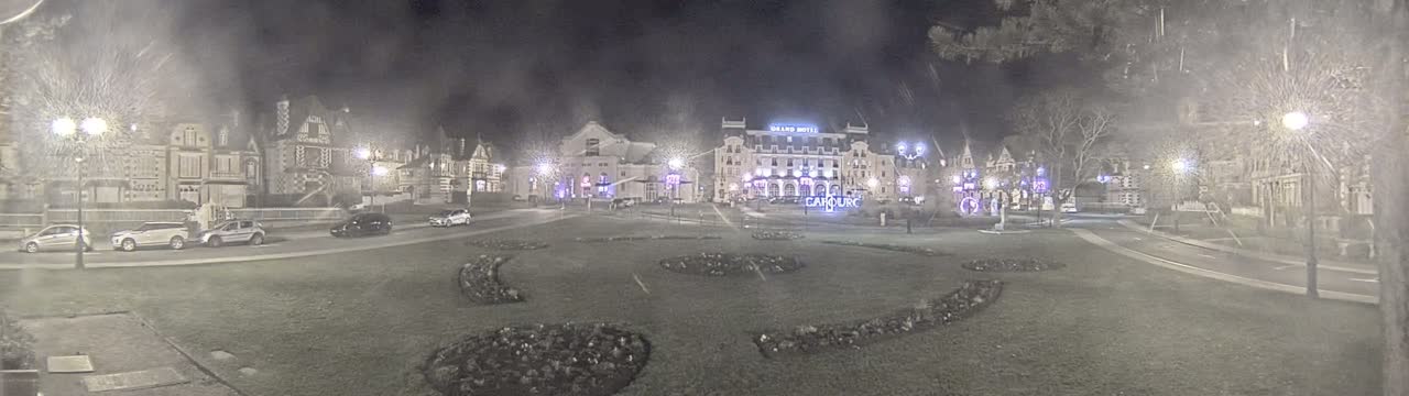A nighttime view of a town square with several buildings, some cars, and decorative landscaping under hazy or snowy conditions.