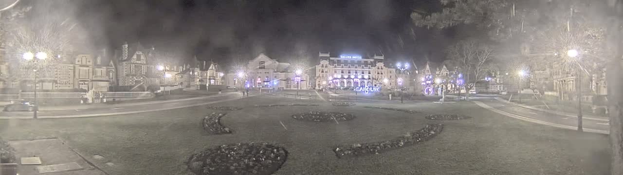 A nighttime view of a town square with several buildings, including a large hotel, illuminated by streetlights in hazy conditions.