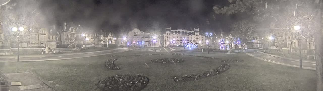 A nighttime scene shows a town square with several buildings, including a large hotel, surrounded by a grassy area with landscaping, under snowy conditions.