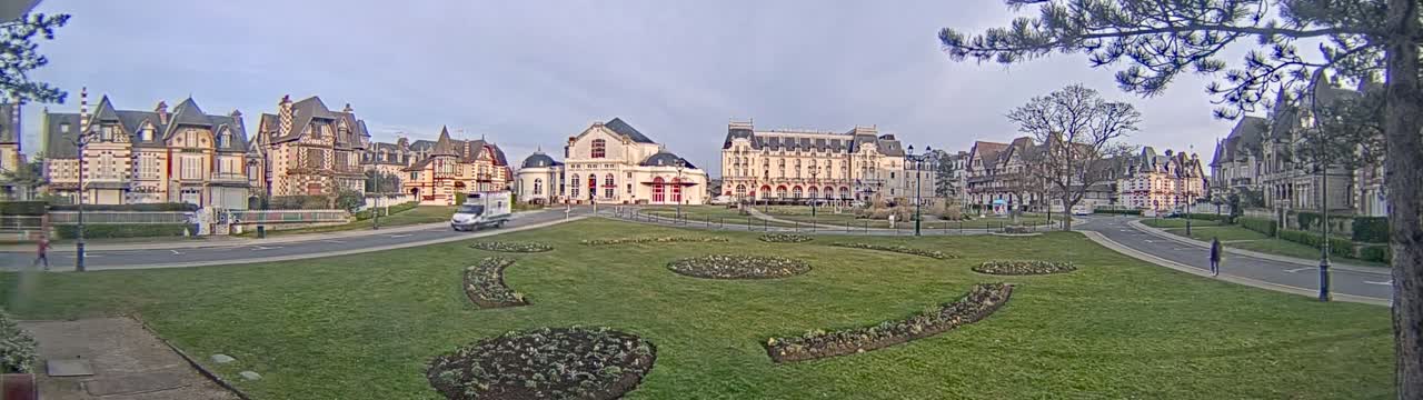 A town square with several large, ornate buildings surrounding a grassy area with landscaped flower beds, under a partly cloudy sky.