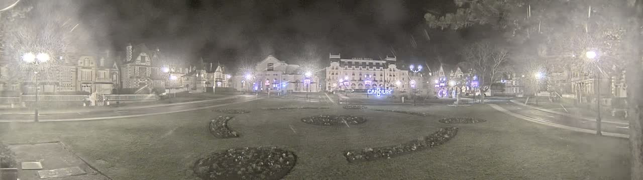 A nighttime view of a town square with several buildings, including a large hotel, surrounded by snow-covered grass and trees under a hazy, snowy sky.