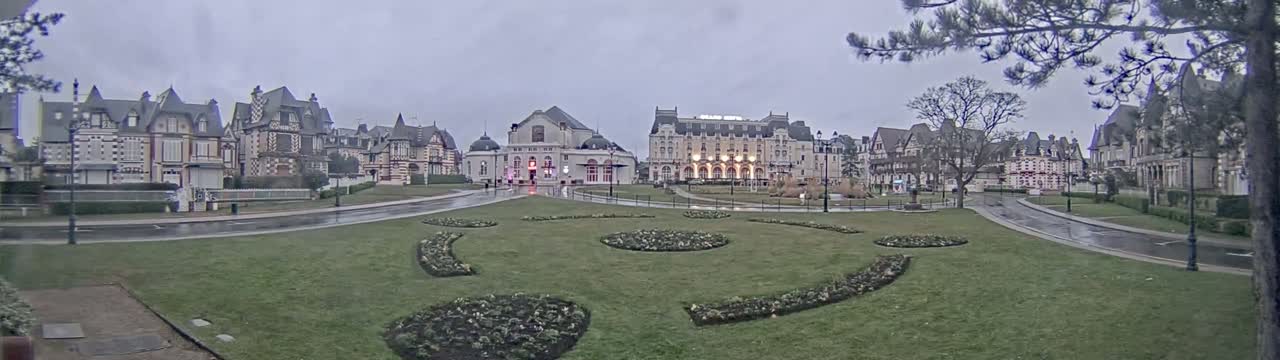 A wet, overcast day reveals a grassy plaza with landscaped flower beds, bordered by several large, ornate buildings.