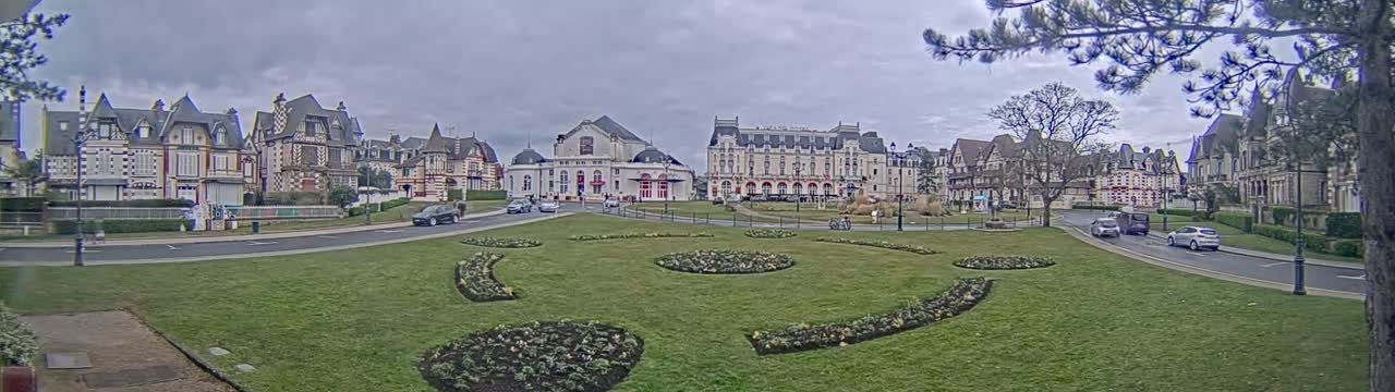 A grassy town square with ornate flowerbeds is surrounded by several large, multi-story buildings under an overcast sky.