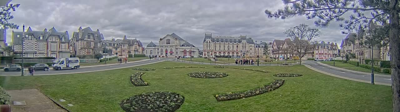 A town square with ornate buildings, flowerbeds, and a few cars is seen under an overcast sky.