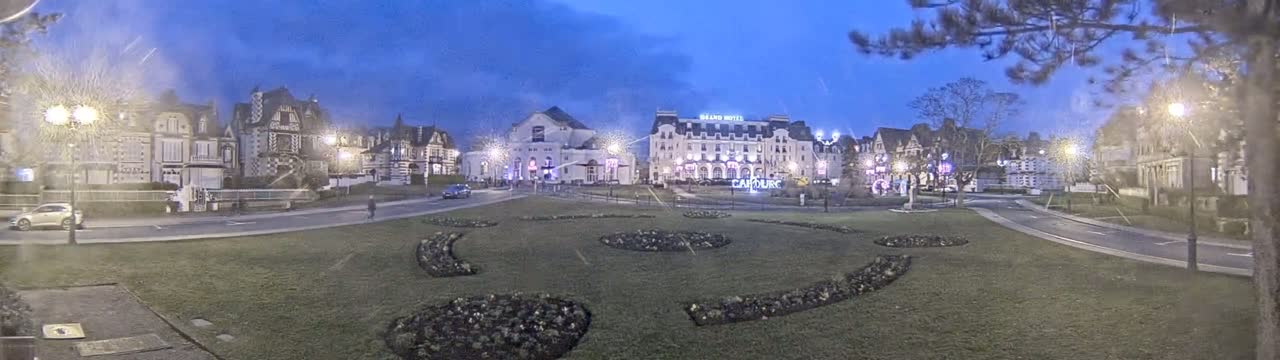 A town square at twilight, with several buildings, including a large hotel, lit up and surrounded by landscaped gardens.