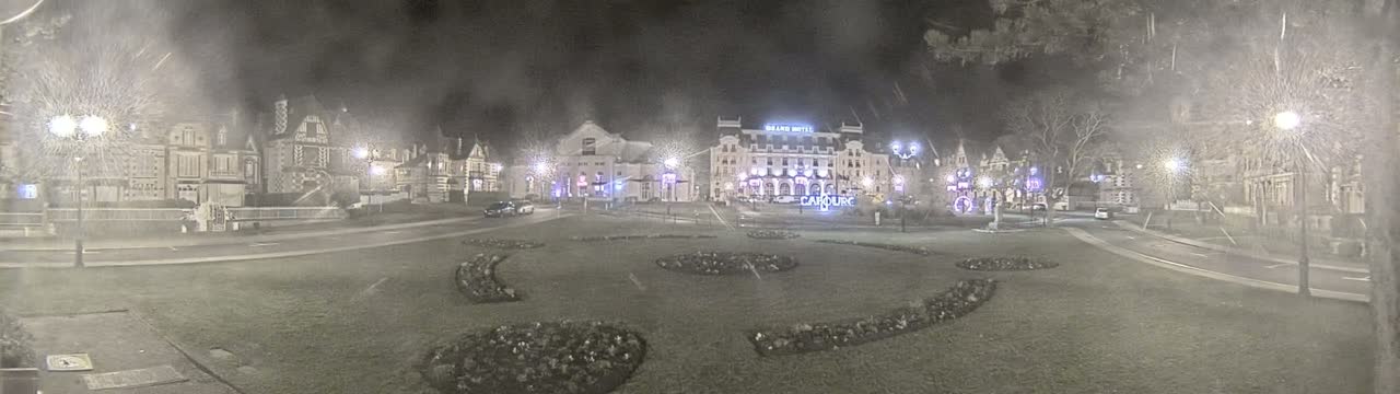 A snow-covered town square at night, illuminated by streetlights, features several buildings, including a large hotel, and landscaped flowerbeds.