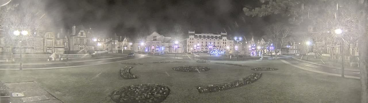 A nighttime, snowy scene shows a town square with several buildings, including a large hotel, surrounded by landscaped areas and illuminated by streetlights.