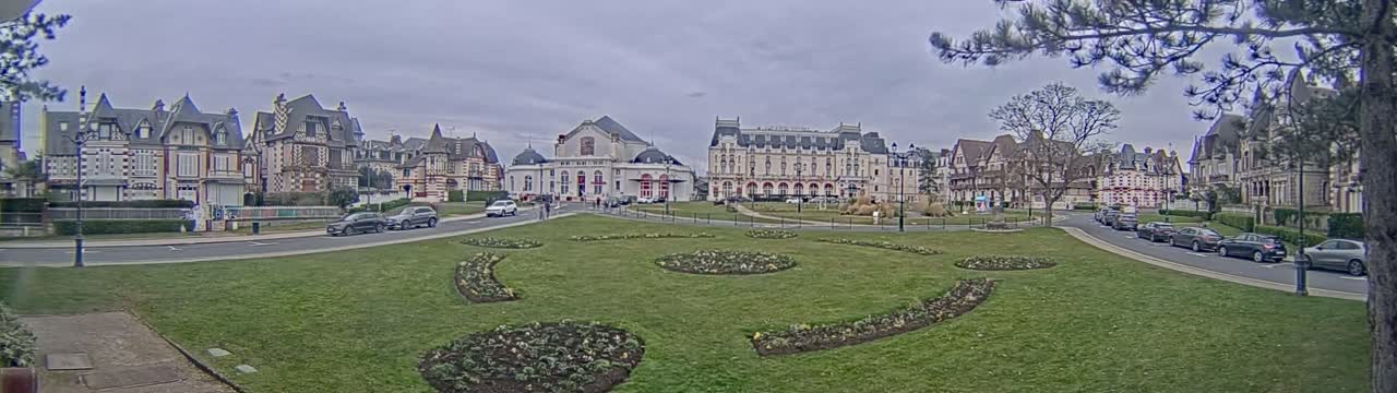A town square with ornate buildings, a grassy area with flowerbeds, and several cars parked along the street on an overcast day.