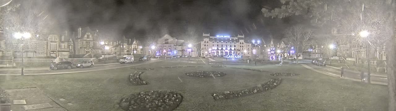 A nighttime, snowy scene shows a town square with several buildings, including a large hotel, and cars parked along the sides of a grassy area with decorative landscaping.