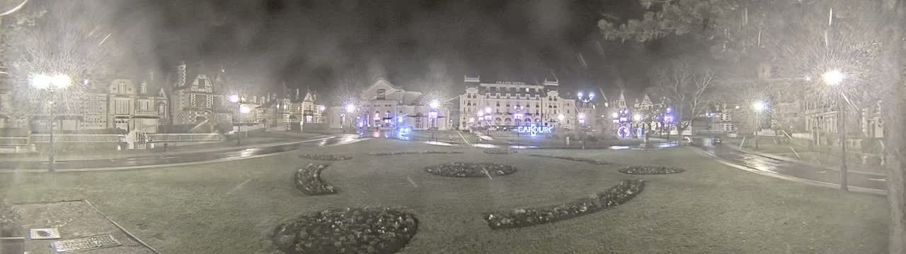 A snow-covered town square at night, featuring several buildings and decorative landscaping illuminated by streetlights.