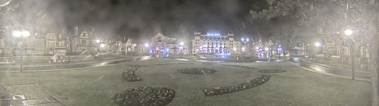 A nighttime scene of a town square with several buildings, landscaped areas, and wet roads under a misty or snowy sky.