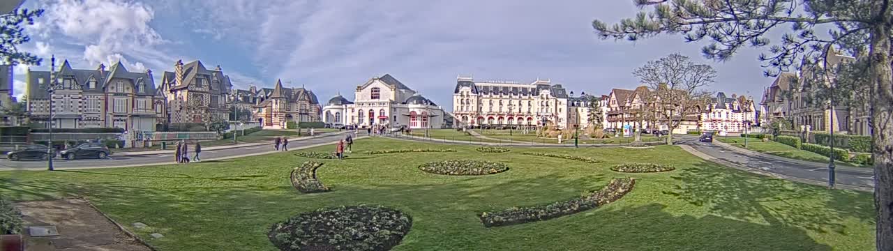 A grassy park with patterned flowerbeds is in front of a row of ornate buildings on a partly sunny day.
