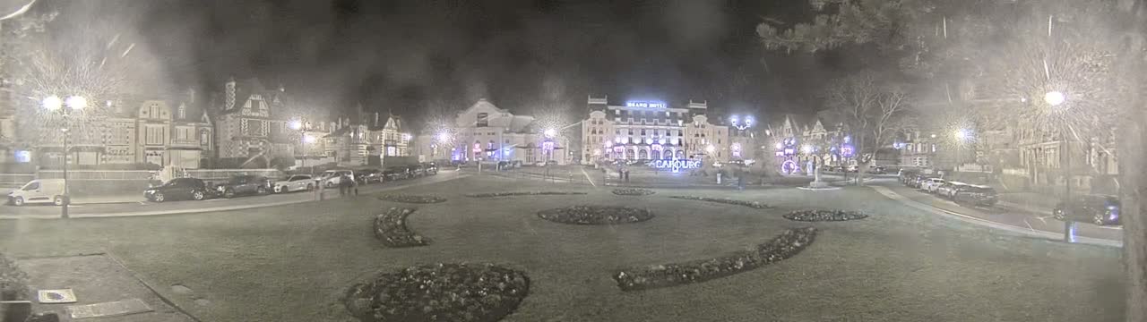 A nighttime scene shows a town square with several buildings, including a large hotel,  illuminated by streetlights and some holiday decorations, with cars parked along the sides and a grassy area in the center.