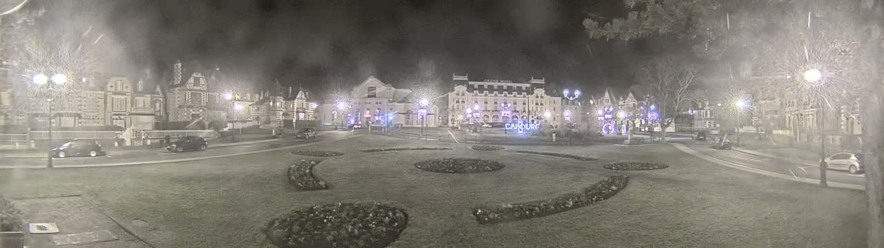 A nighttime scene shows several buildings, including a large hotel, surrounded by a grassy area with landscaped flower beds, and sparsely parked cars under illuminated streetlights.