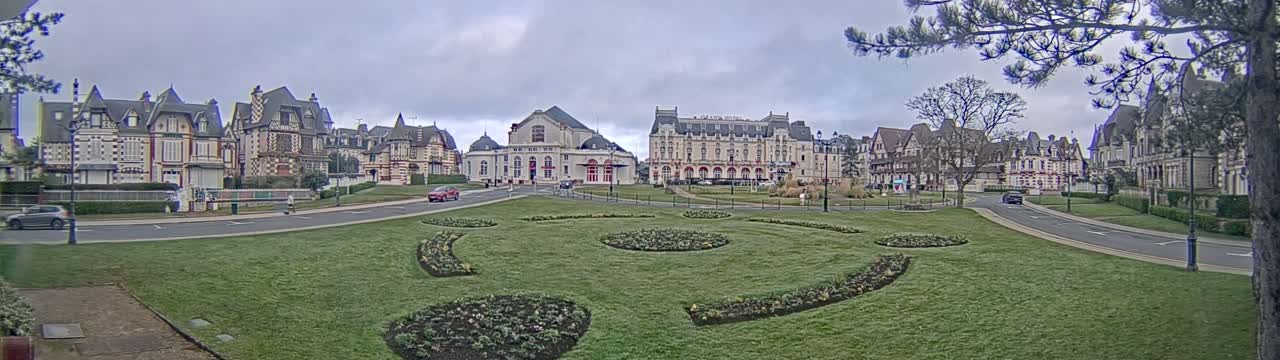 A row of ornate buildings stands across a grassy park with flowerbeds under an overcast sky.
