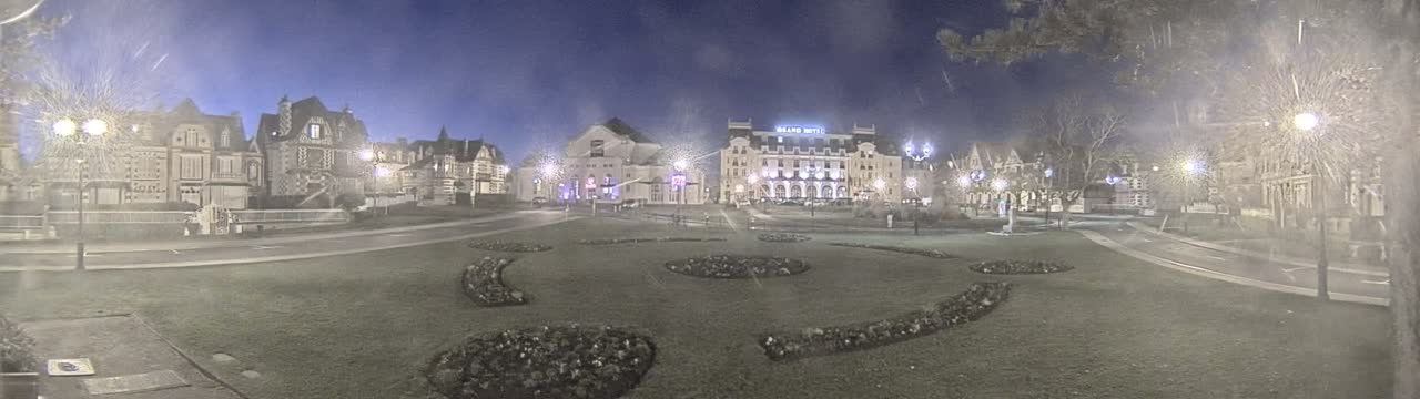 A town square at night in foggy conditions, featuring several ornate buildings, including a hotel, and landscaped flower beds.