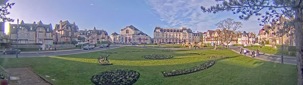 A grassy park with decorative flowerbeds is in front of a row of ornate buildings under a mostly sunny sky.