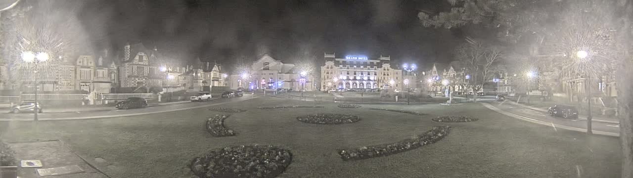 A nighttime view of a town square with several buildings, including a large hotel, and landscaped areas, under hazy or snowy conditions.