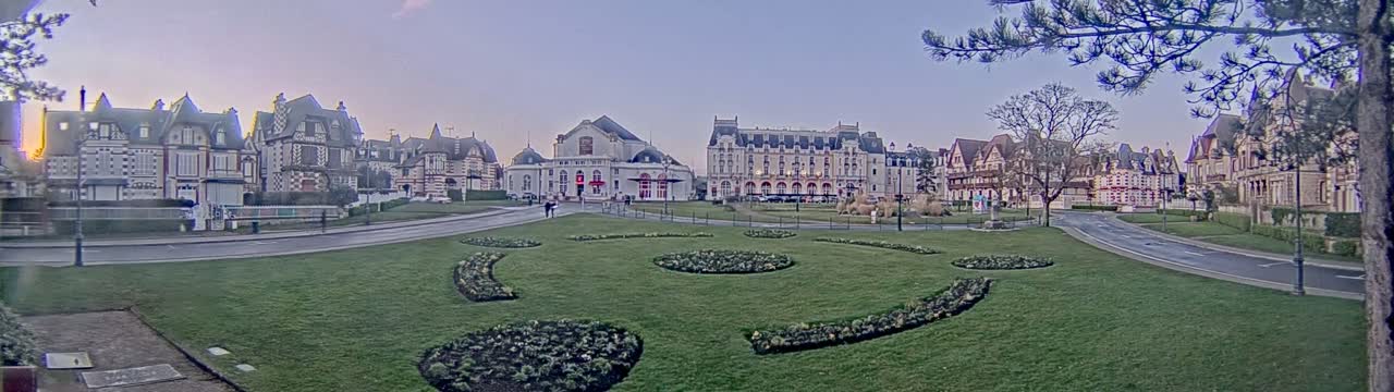 A wide, grassy park with patterned flowerbeds is situated in front of a row of ornate buildings under a twilight sky.