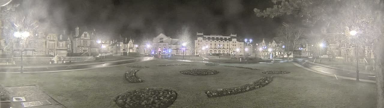 A nighttime scene of a town square with several buildings, some of which are lit, surrounded by a grassy area with landscaped flower beds, under hazy or snowy conditions.