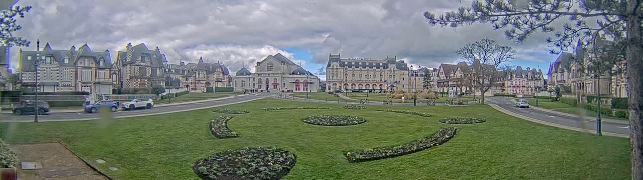 A wide, grassy plaza with patterned flowerbeds is centered between several large, ornate buildings under a cloudy sky.