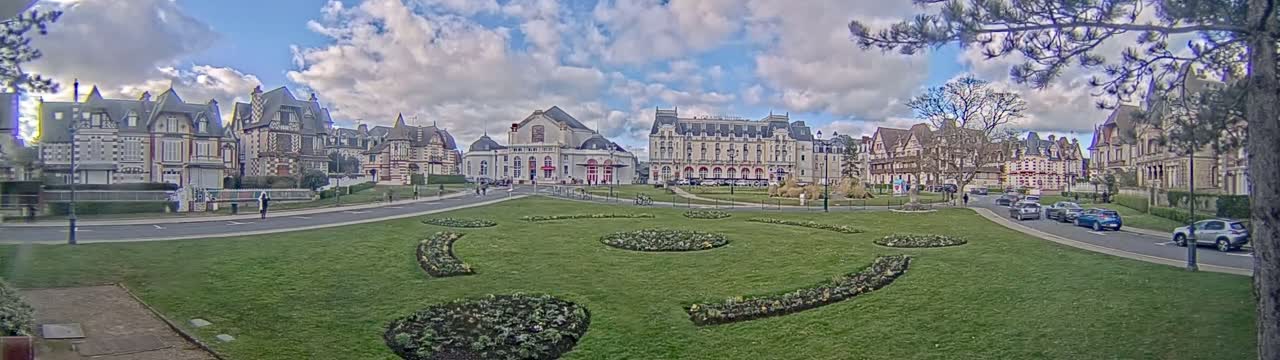 A grassy town square with ornate buildings, including a large hotel and theater, surrounds it under a partly cloudy sky.