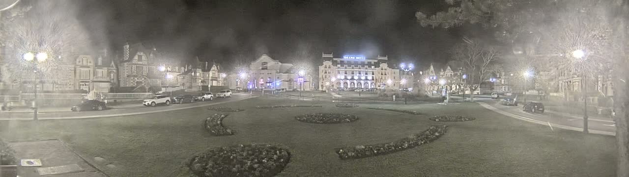 A nighttime scene of a town square with several buildings, cars, and decorative landscaping, under hazy or snowy conditions.