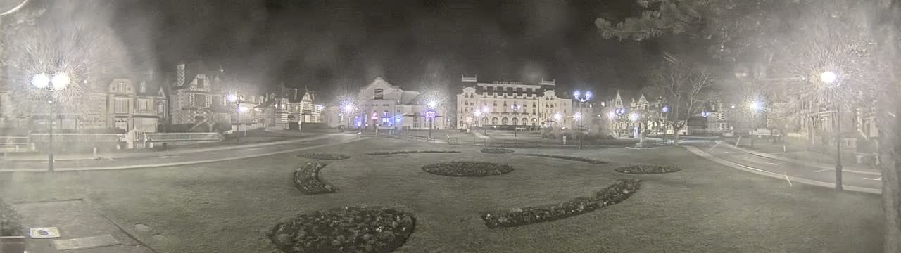 A snow-covered town square at night, with several buildings illuminated by streetlights and landscaping features visible in the foreground.