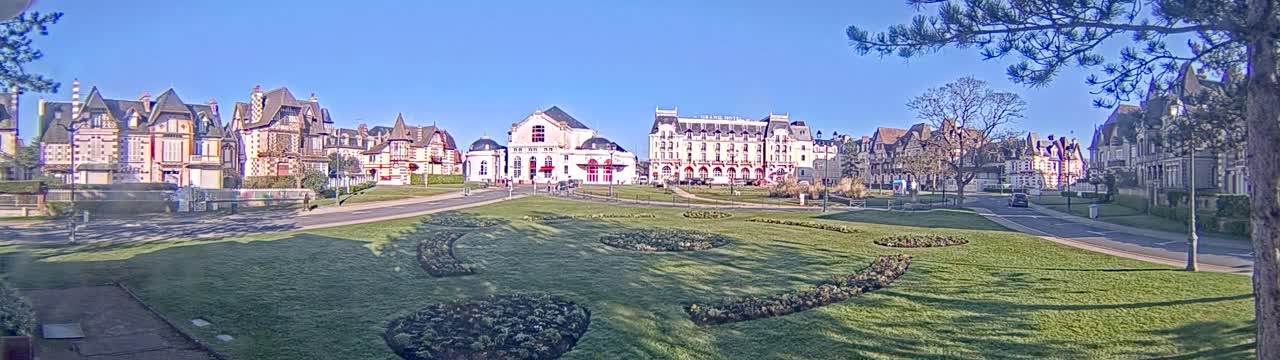 A row of elegant buildings, including a large hotel, sits across a grassy park under a clear, sunny sky.