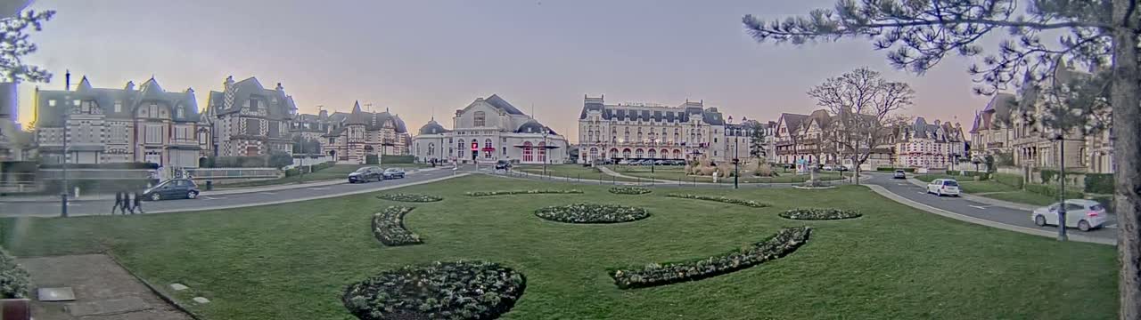 A grassy square with ornate flowerbeds is situated in front of several large, ornate buildings under a pale, dusky sky.