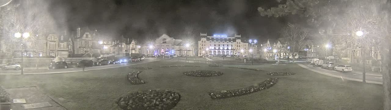 A nighttime scene shows a town square with several buildings, cars parked along the sides, and landscaped flower beds in the center, under hazy or snowy conditions.