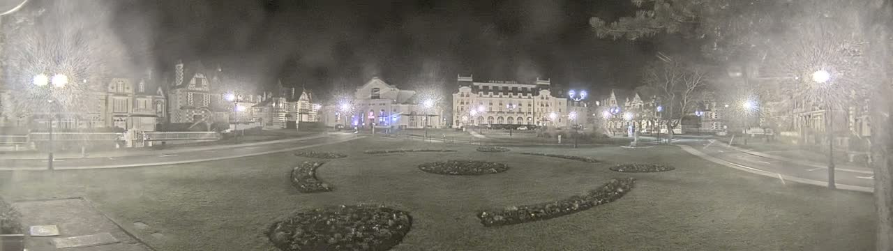 A nighttime view of a town square with several buildings, including a large hotel, surrounded by manicured lawns and illuminated by streetlights in foggy conditions.
