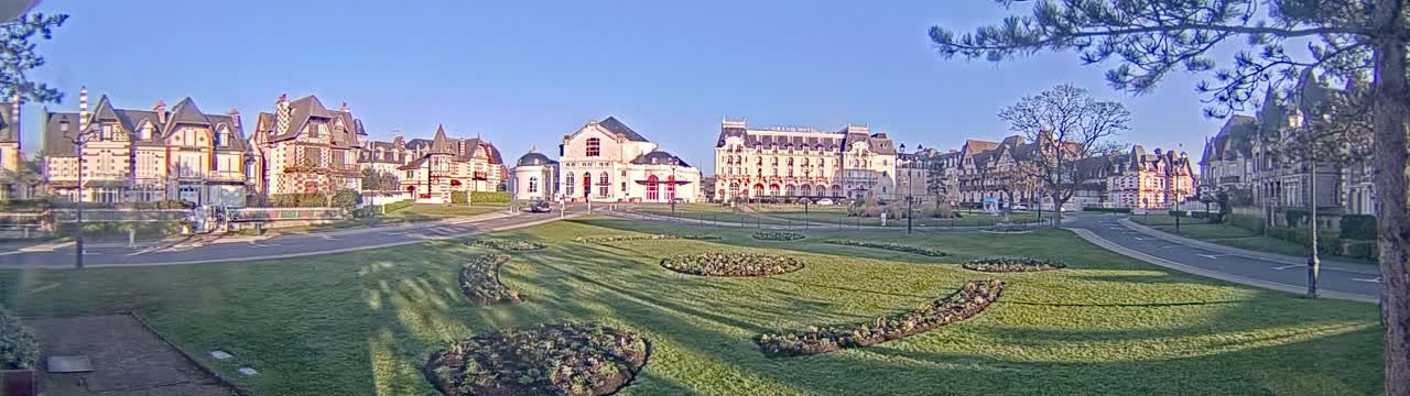 A sunny day reveals a row of elegant buildings, including a large hotel, situated across a grassy park with meticulously landscaped flowerbeds.
