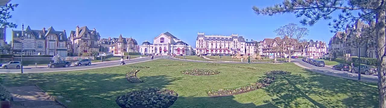 A sunny day reveals a row of elegant buildings and a grassy park with meticulously landscaped flowerbeds, cars parked along the street.