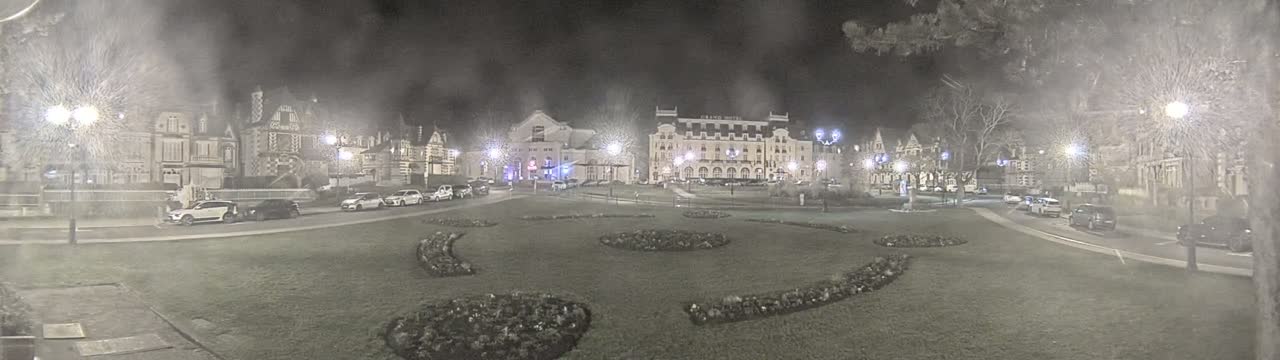 A snow-covered town square at night, featuring several buildings, parked cars, and ornamental landscaping.