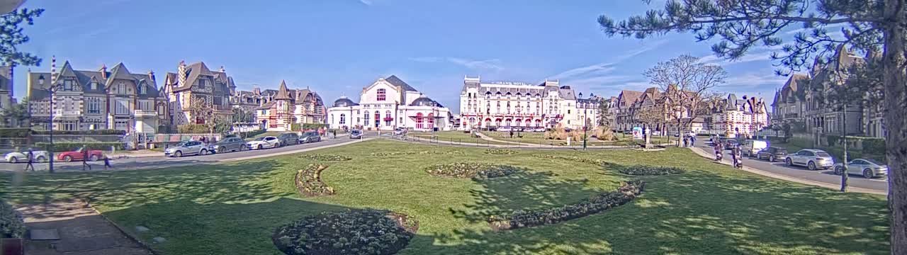A sunny day reveals a town square with several multi-story buildings, cars parked along the road, and a grassy area with landscaping.