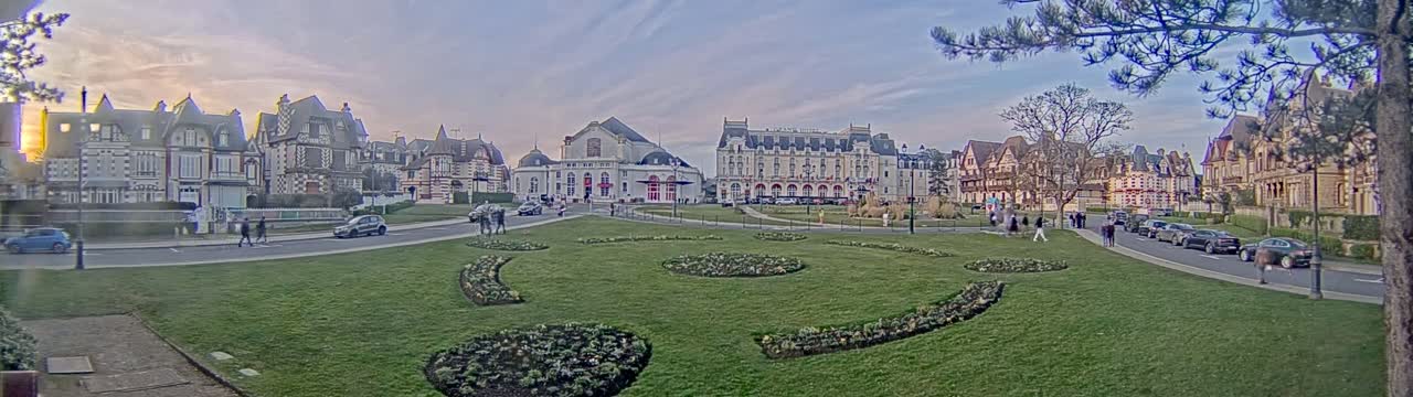 A grassy square with flowerbeds is surrounded by large, ornate buildings under a partly cloudy, sunset sky.