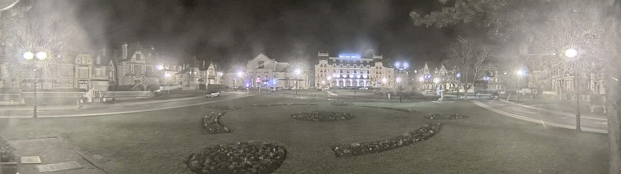 A snow-covered town square at night, illuminated by streetlights, features several buildings including a large hotel, and landscaped areas with low shrubs.