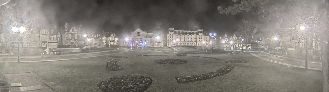 A nighttime view of a town square with several buildings, including a large hotel, surrounded by landscaped gardens under hazy conditions.