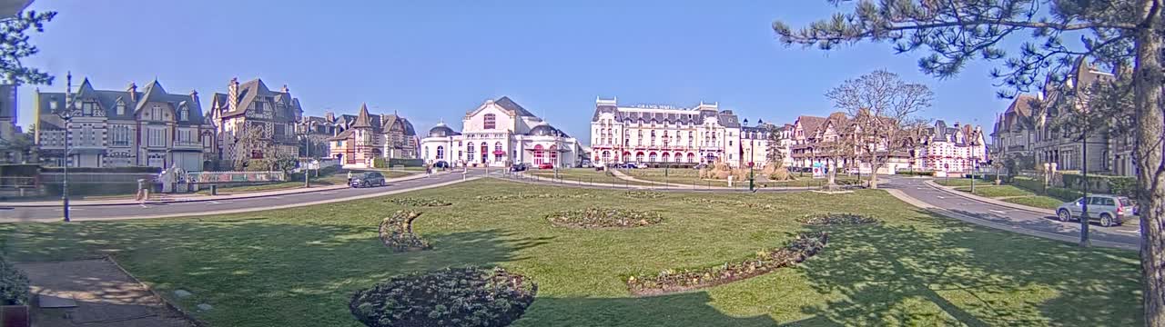 A grassy square with flowerbeds is surrounded by several large, ornate buildings under a clear, sunny sky.